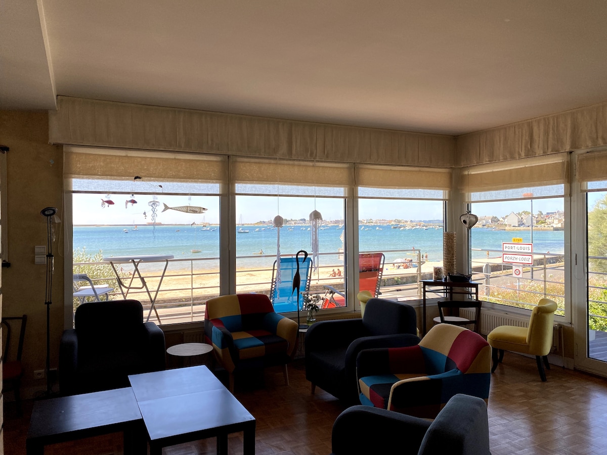 A spacious living area featuring large windows that provide a clear view of the Petite Mer de Gâvres. Colorful armchairs and tables are arranged for relaxation, allowing plenty of natural light to fill the room.