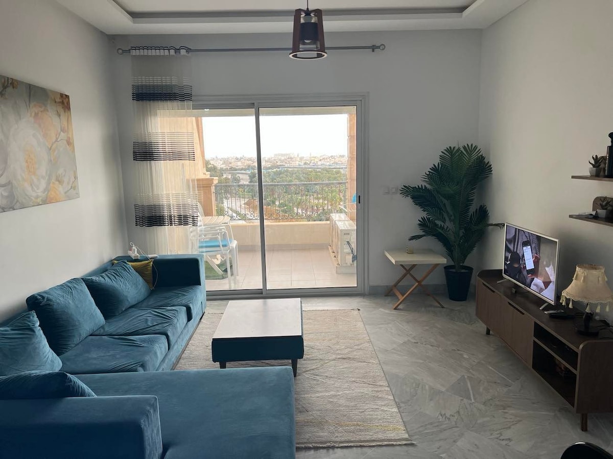 A comfortable living area features a plush blue sofa, a wooden coffee table, and a television on a low cabinet. Natural light floods in through glass sliding doors that lead to a balcony with city views. A small plant adds a touch of greenery to the space.