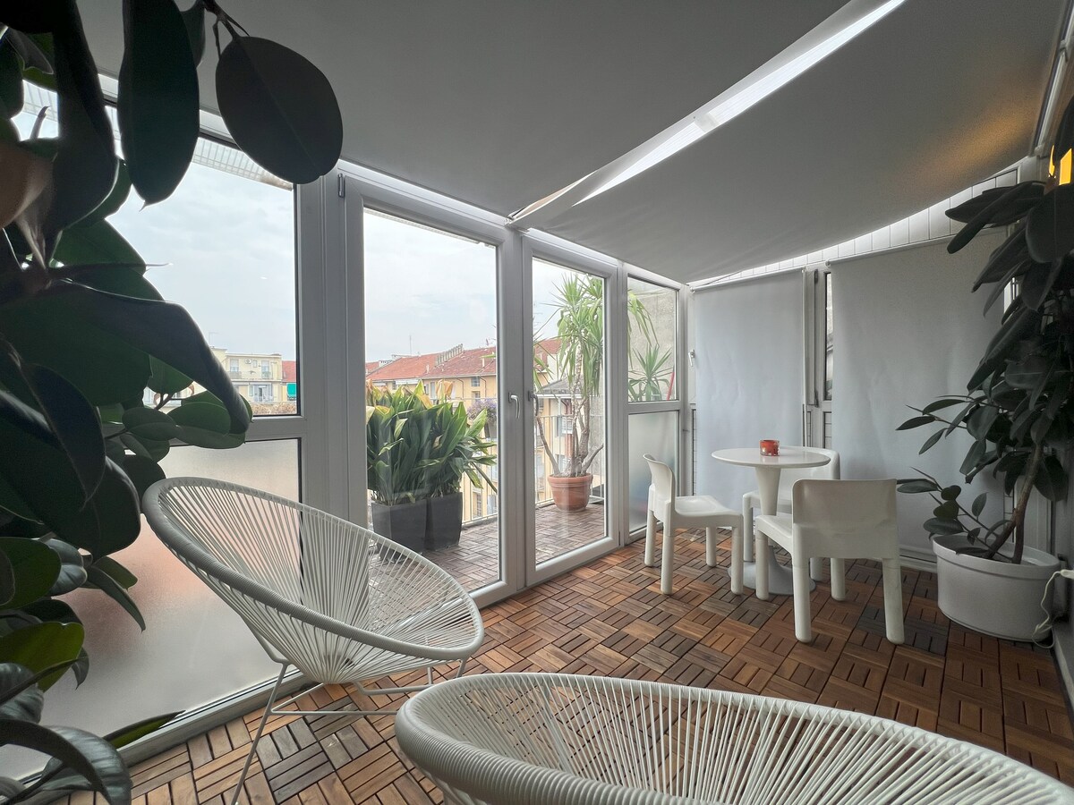 A bright terrace is enclosed by large windows, offering views of surrounding greenery and residential buildings. White chairs surround a round table, with potted plants adding natural elements to the space. The flooring features wooden tiles arranged in a patterned layout.