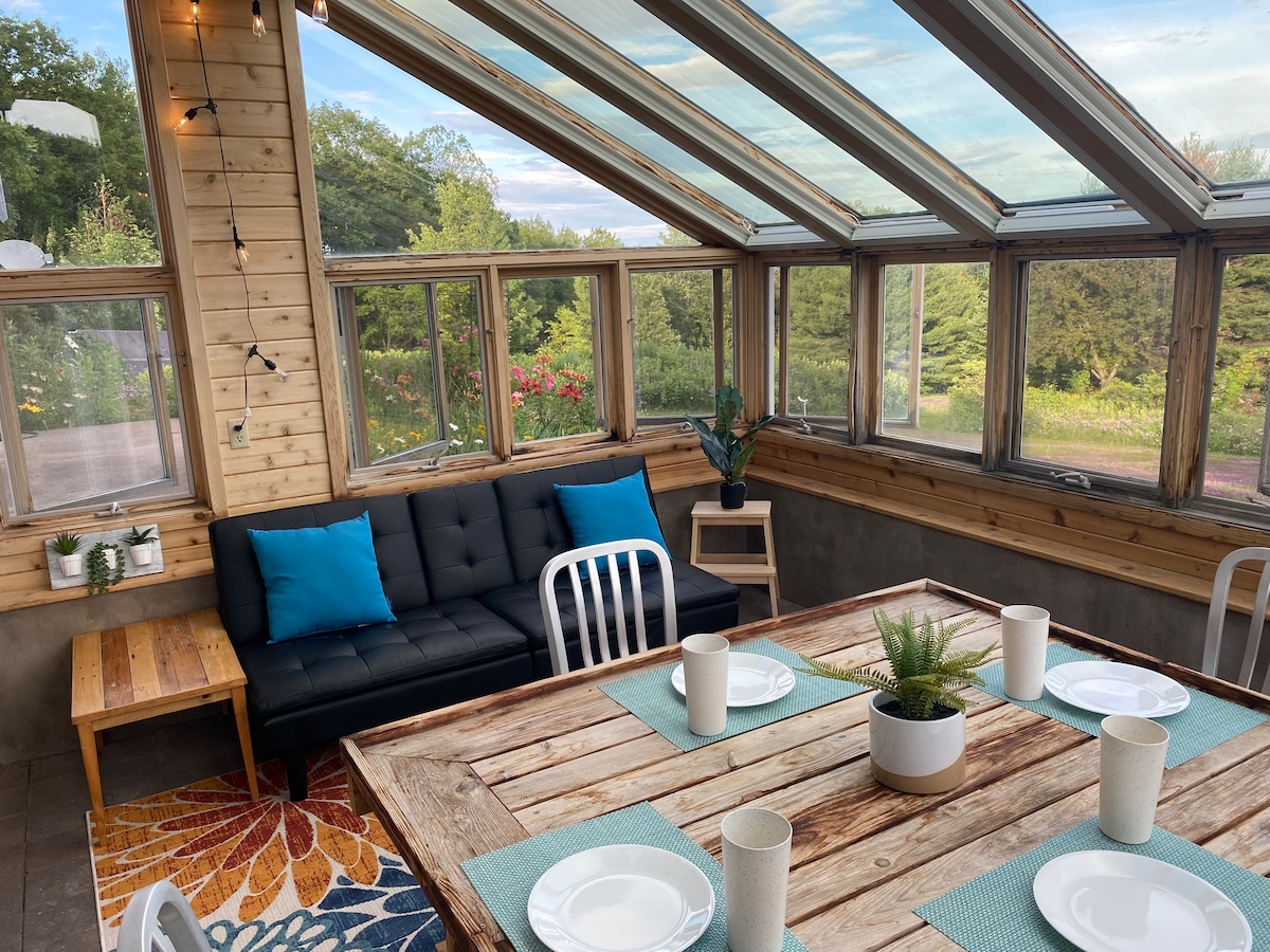 A sunlit space features a wooden dining table set with plates and cups, surrounded by chairs. A comfortable sofa with blue cushions is positioned nearby. Large glass panels illuminate the room, framed by natural wood accents and lush outdoor views.