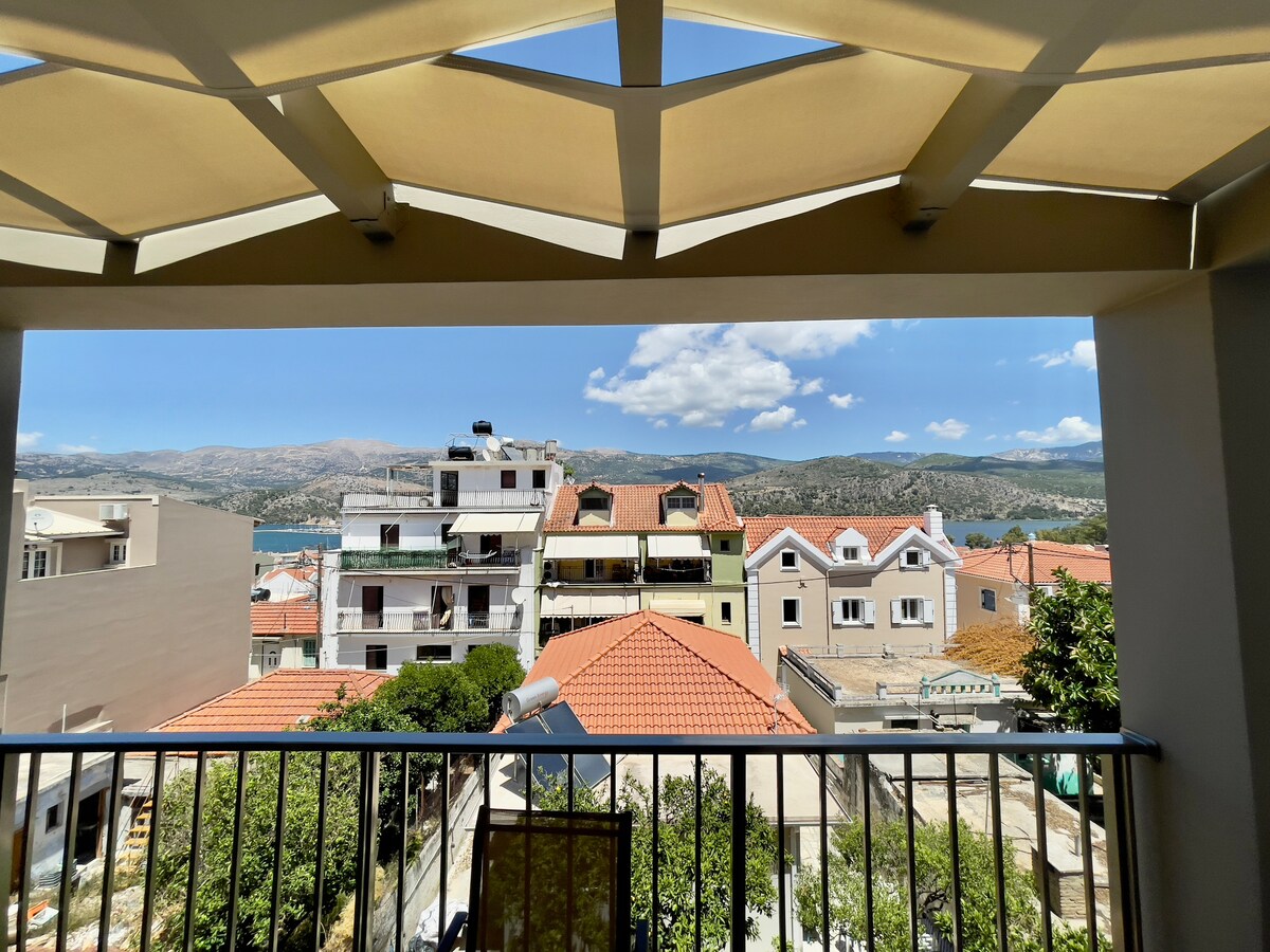 A view from the balcony showcases a mix of residential buildings with terracotta roofs and a backdrop of rolling hills. The sky is bright blue with scattered clouds, providing a clear perspective of the surrounding area. Sunlight filters through a canopy above.