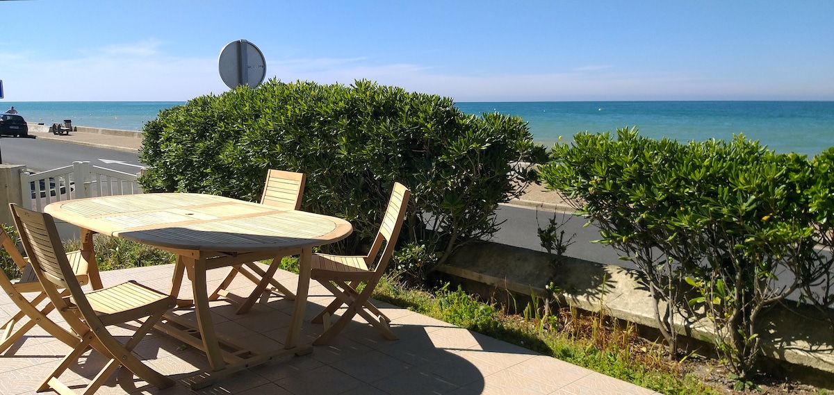 A round wooden table with several folding chairs is situated on a terrace, surrounded by lush greenery. The view extends to a sandy beach and the ocean, with a clear blue sky framing the scene.