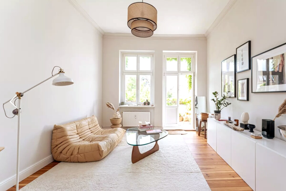 A bright living room is shown, featuring a low, tan-colored sofa and a glass coffee table. Two large windows allow natural light to illuminate the space, complemented by plants and framed artwork on the walls. A soft area rug covers the wooden floor.
