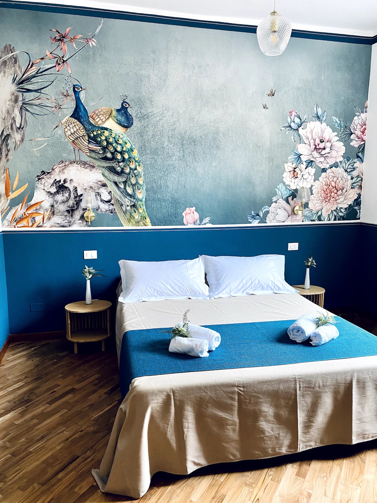 A harmonious bedroom features a spacious bed covered with a light beige bedspread. Decorative elements include a vibrant mural of peacocks and floral designs on the wall, complemented by a rich blue accent wall. Two bedside tables hold small vases with fresh flowers.