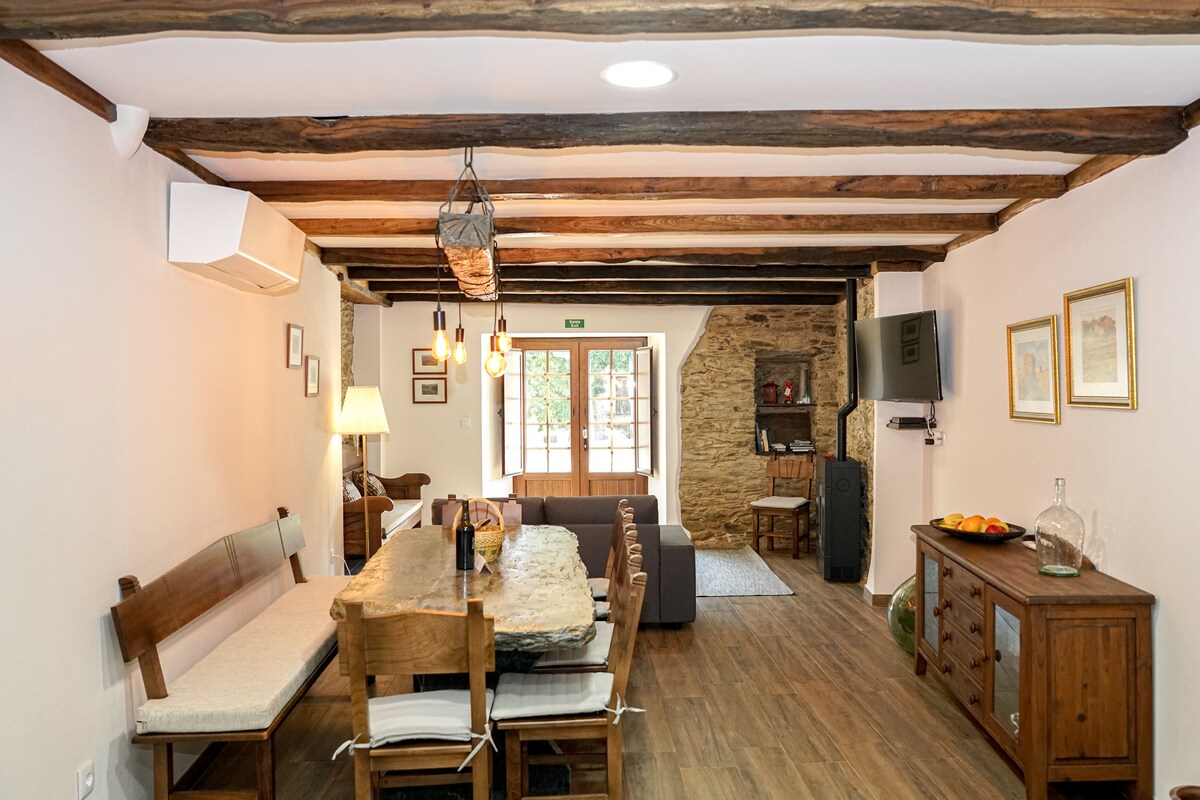 An open living space showcases exposed wooden beams and a rustic wooden dining table surrounded by matching chairs. A cozy seating area is visible, featuring a grey sofa and a stone wall that adds character. Natural light streams through the windows, enhancing the ambiance.