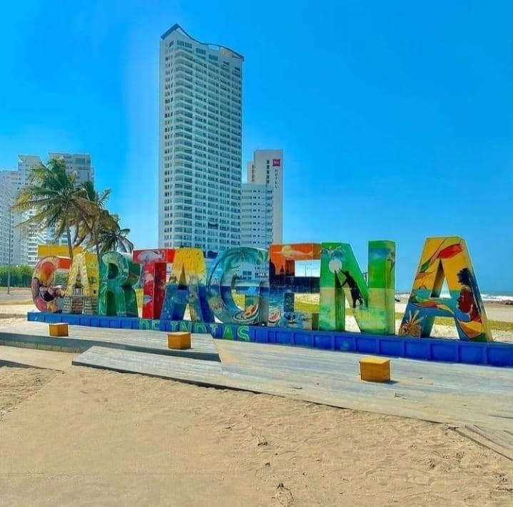 A vibrant sign spelling 'CARTAGENA' is displayed on the beach, with colorful letters adorned with artistic designs. Behind the sign, tall buildings rise against a clear blue sky, while palm trees sway gently in the breeze, enhancing the coastal atmosphere.