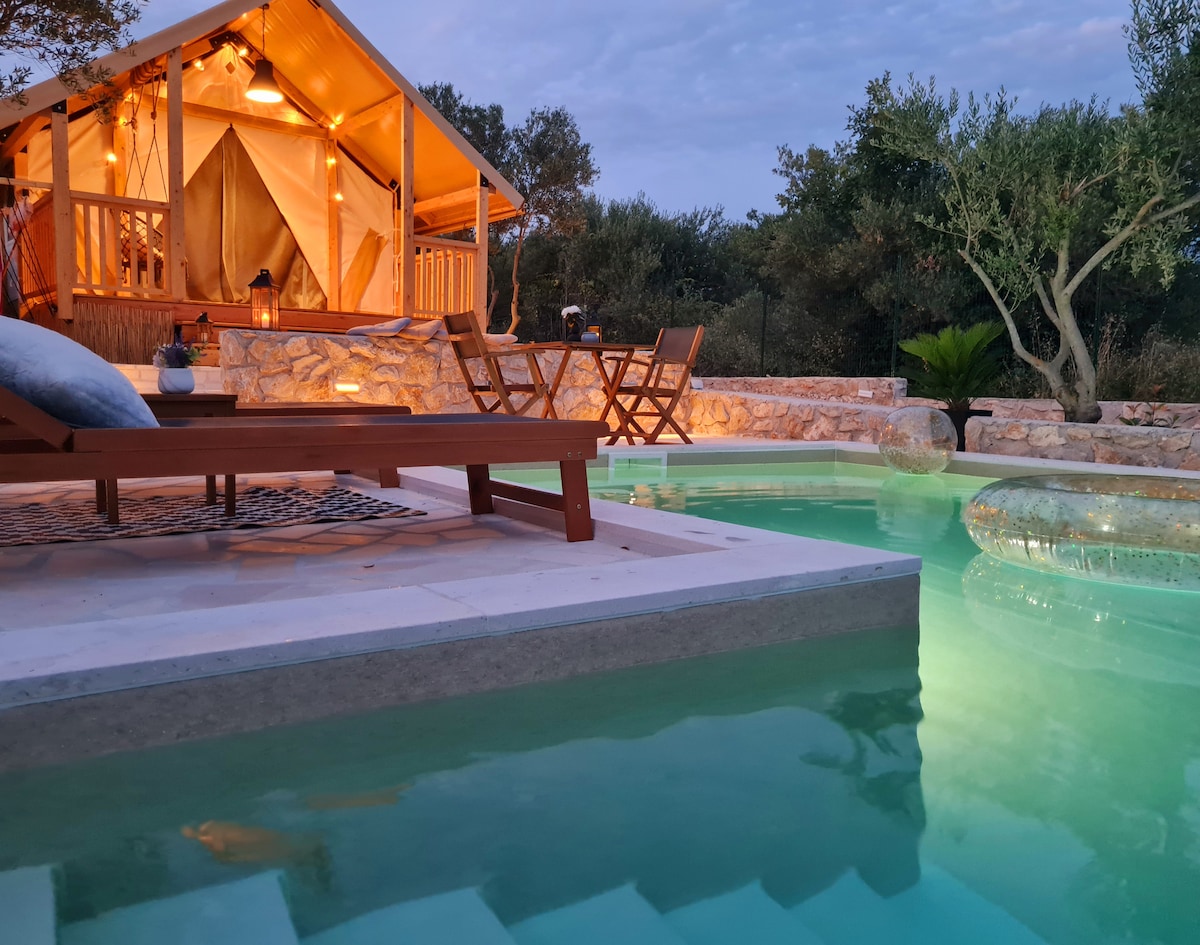 A private pool is visible at the forefront, featuring steps leading into turquoise water. Adjacent to the pool, a wooden table with two chairs is set, complemented by a decorative plant. The luxury tent, with lit interiors, is nestled under olive trees, enhancing the serene environment.