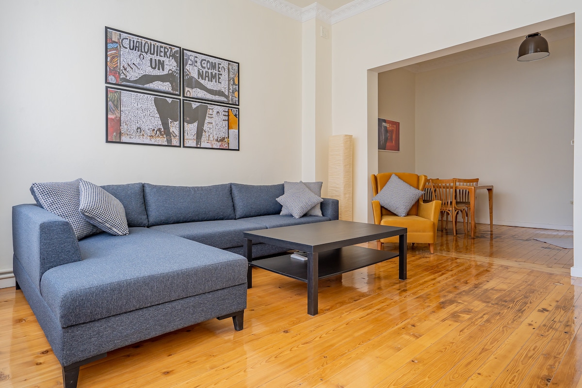 A spacious living area is highlighted with a large 'L' shaped sofa, accompanied by decorative cushions. A coffee table sits in front, and artwork adorns the wall above. The dining area is visible through the open space, featuring a wooden table and chairs.