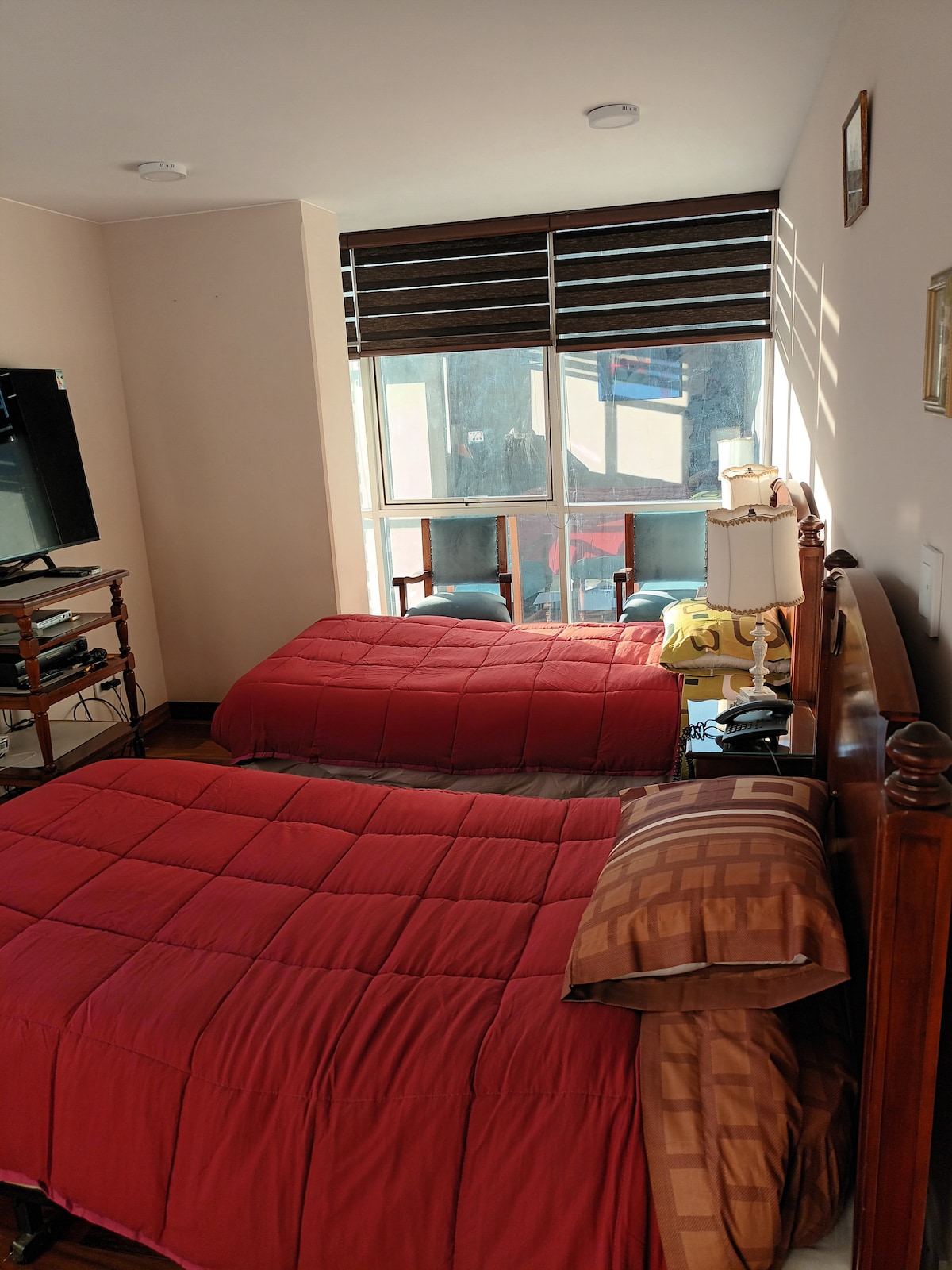 A well-lit bedroom features two neatly made beds with burgundy comforters. A wooden bedside table holds a lamp, and a television is positioned across from the beds. Natural light enters through a window with horizontal blinds, illuminating the space.