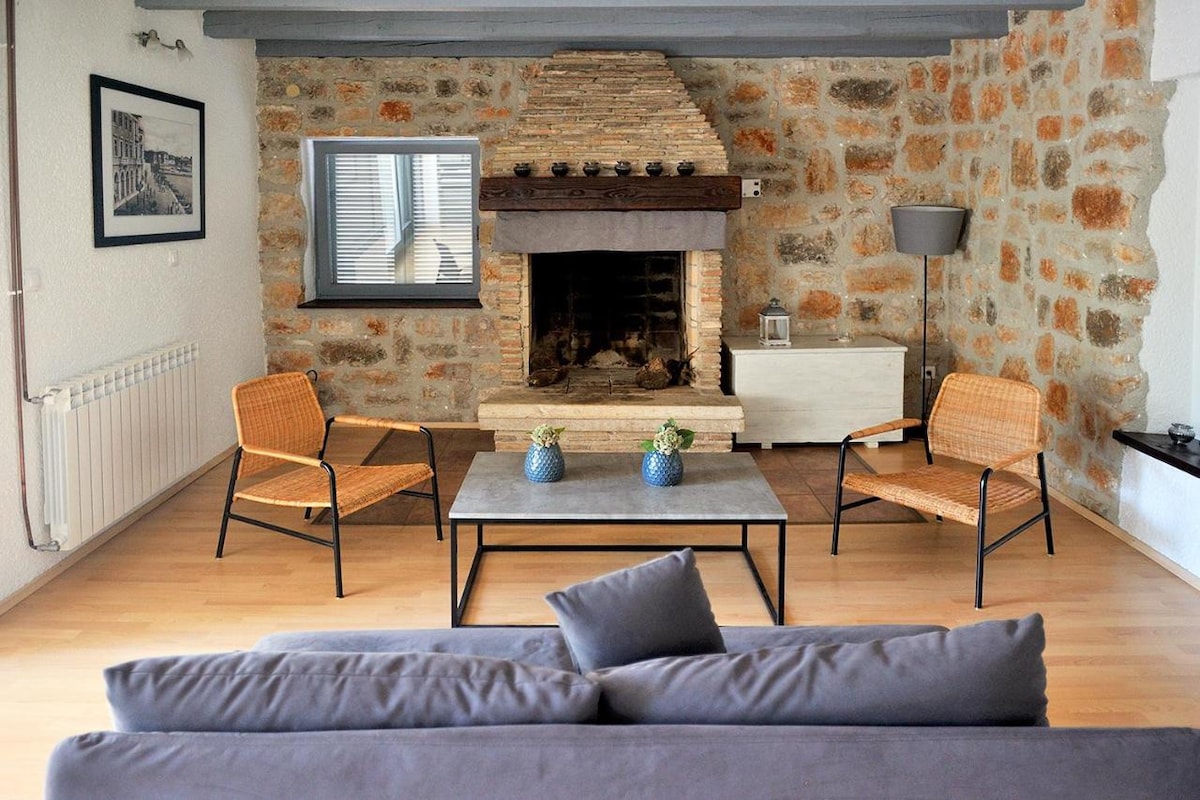 A cozy living space features a stone fireplace as a central element. Two woven chairs are positioned around a compact table, decorated with small potted plants. A soft sofa is visible in the foreground, while a large window allows natural light to brighten the room.