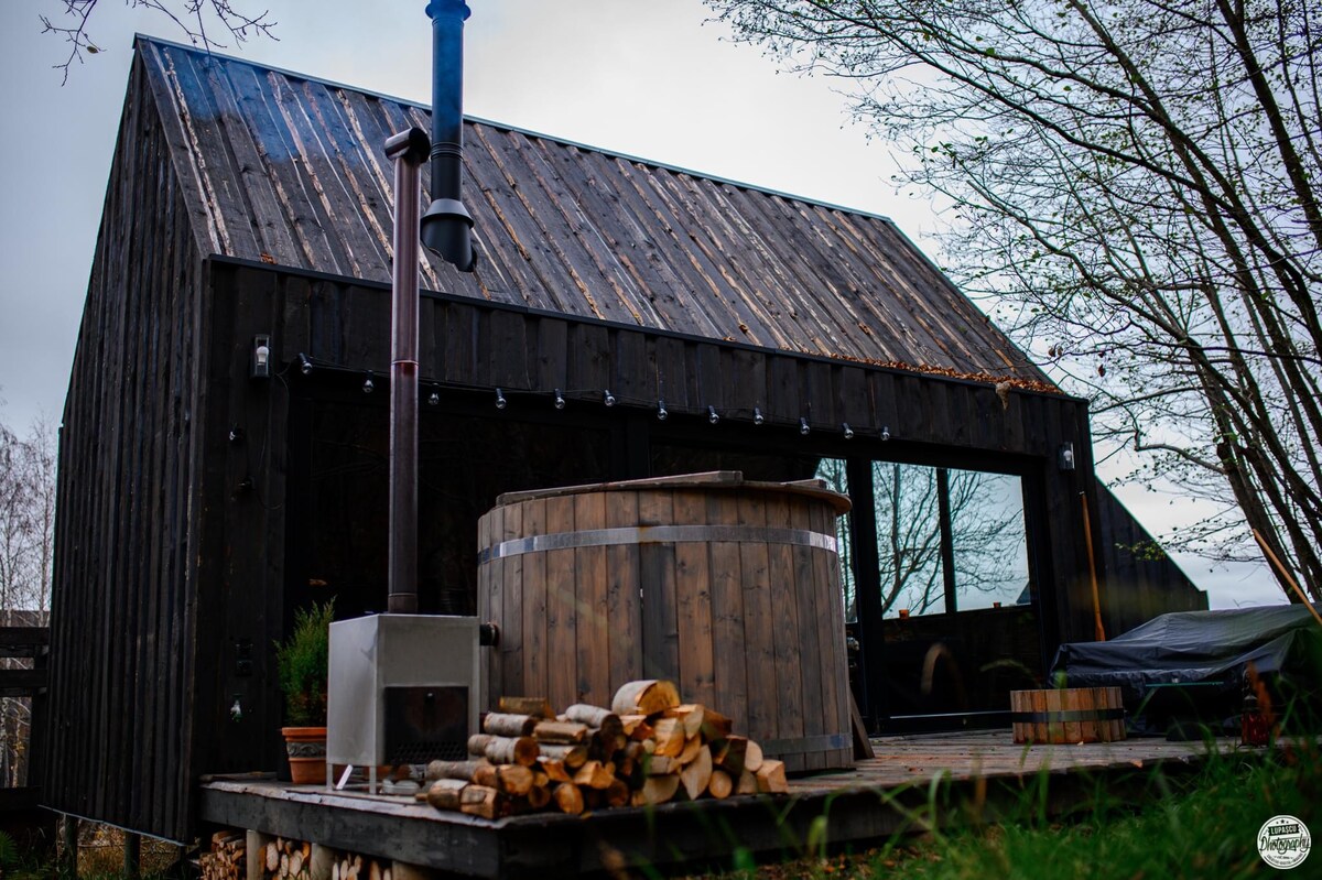 A wooden structure with a sloped roof features large windows that invite natural light. In front, a circular hot tub made of wood is located on a raised deck, accompanied by a stack of firewood nearby. Surrounding greenery enhances the serene atmosphere.