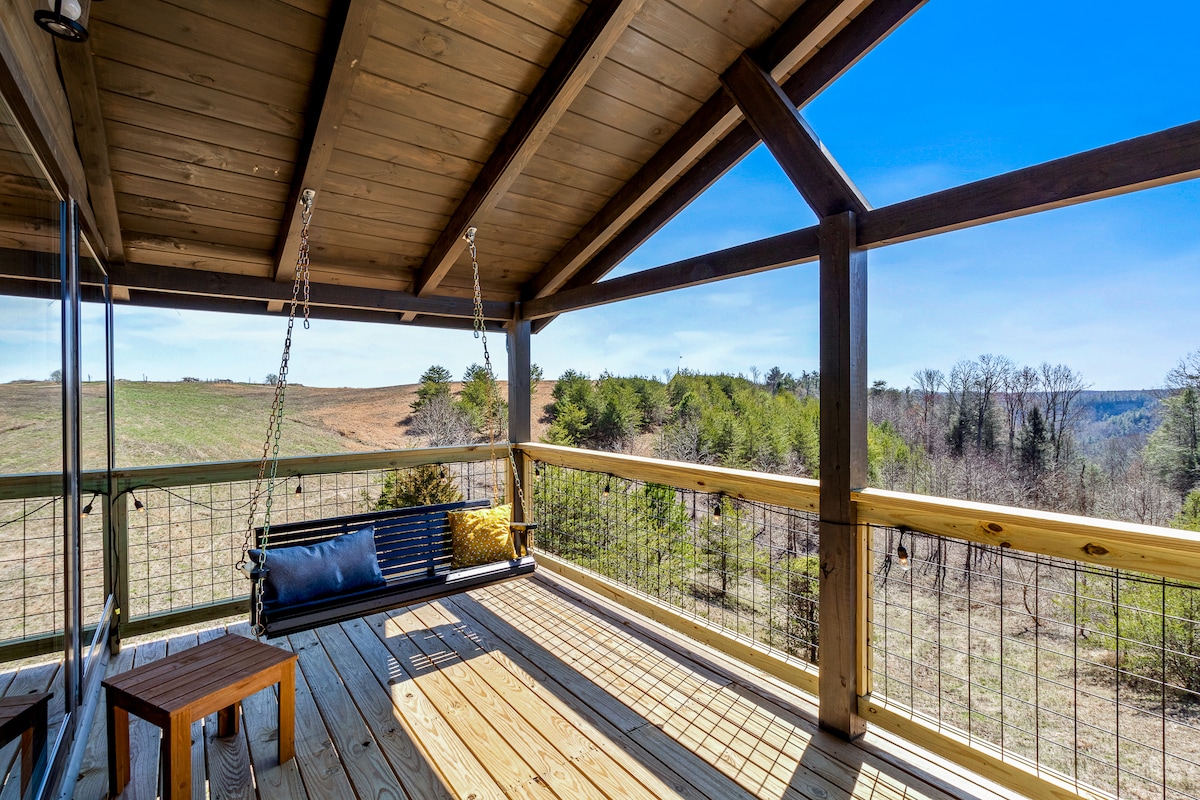 An outdoor deck is shown, featuring a wooden swing seat with a yellow pillow. The area is designed with a natural wood finish, allowing for panoramic views of the lush green landscape and distant rolling hills under a clear blue sky.