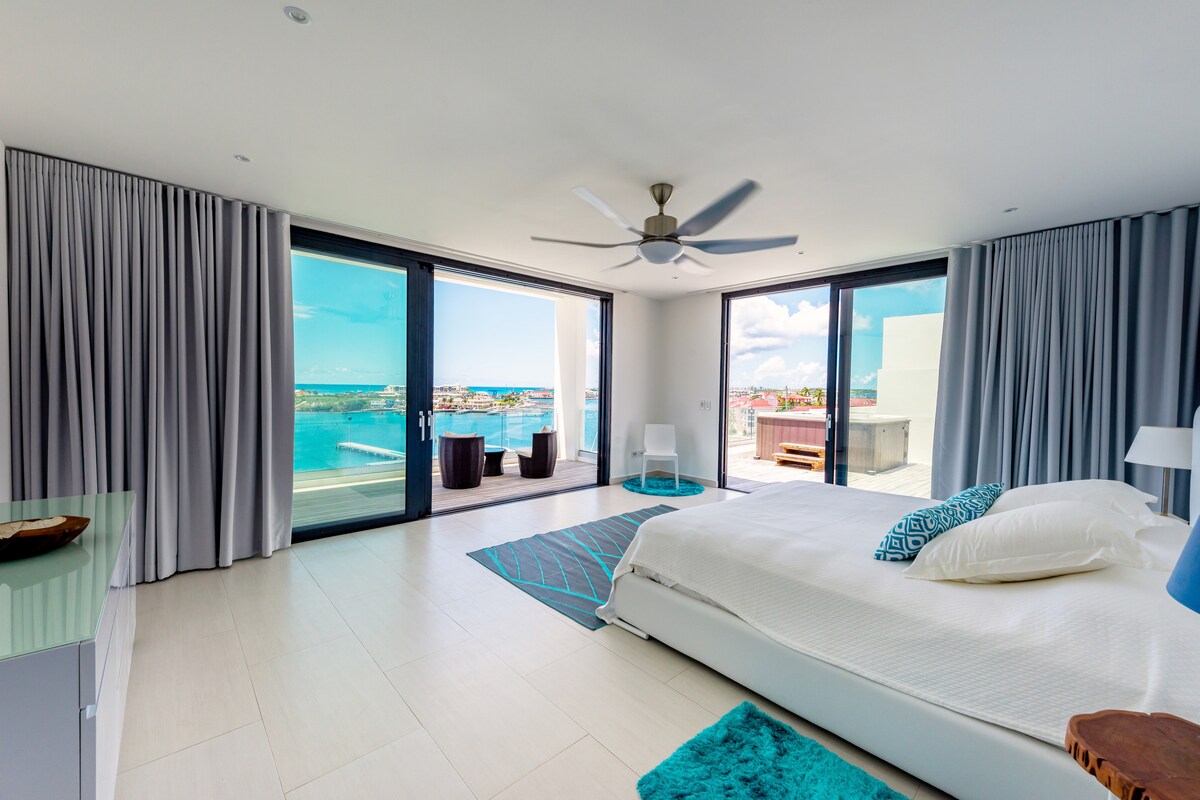A spacious bedroom features large glass doors that open to a deck with a panoramic view of the lagoon and marina. Light-colored walls and soft furnishings provide a calm ambiance, while a ceiling fan enhances air circulation. A plush area rug complements the overall design.