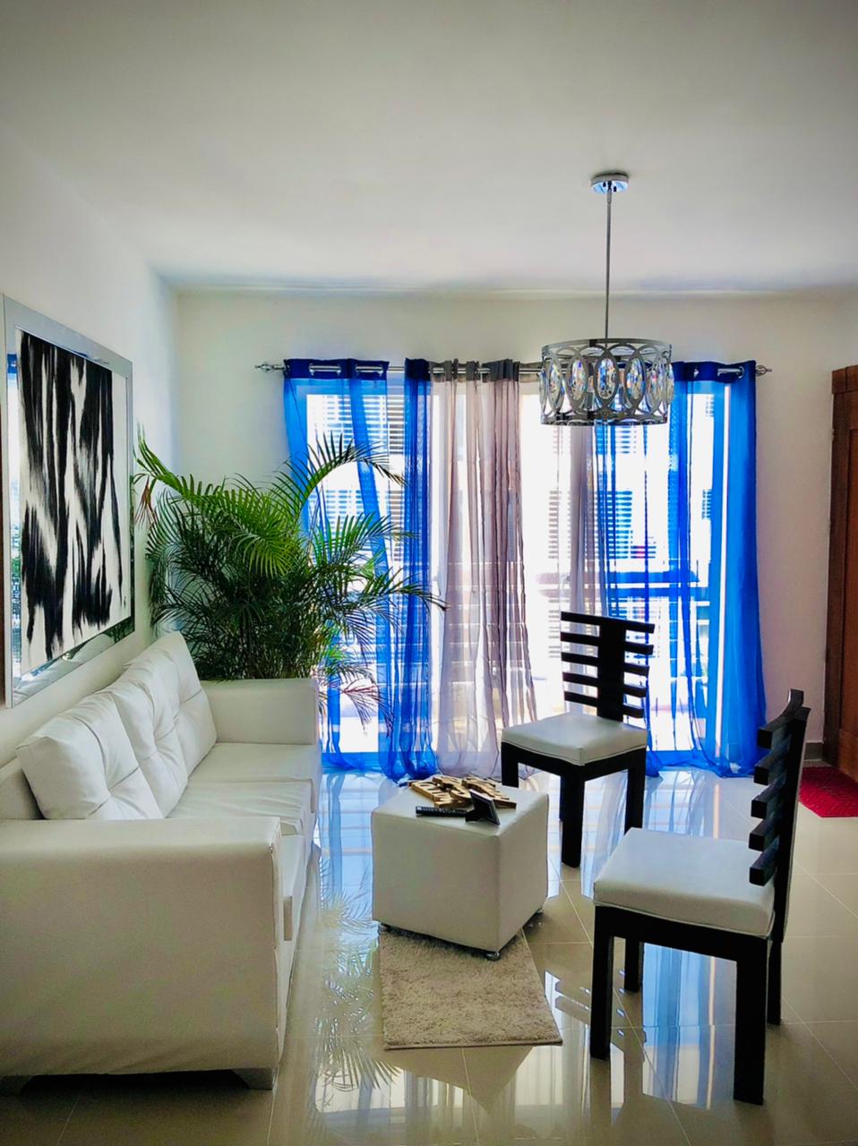 A bright living area is showcased with a white sofa and two black chairs around a small white ottoman. Large windows are adorned with sheer and dark curtains, allowing natural light to illuminate the space. A green plant adds a touch of nature to the room.
