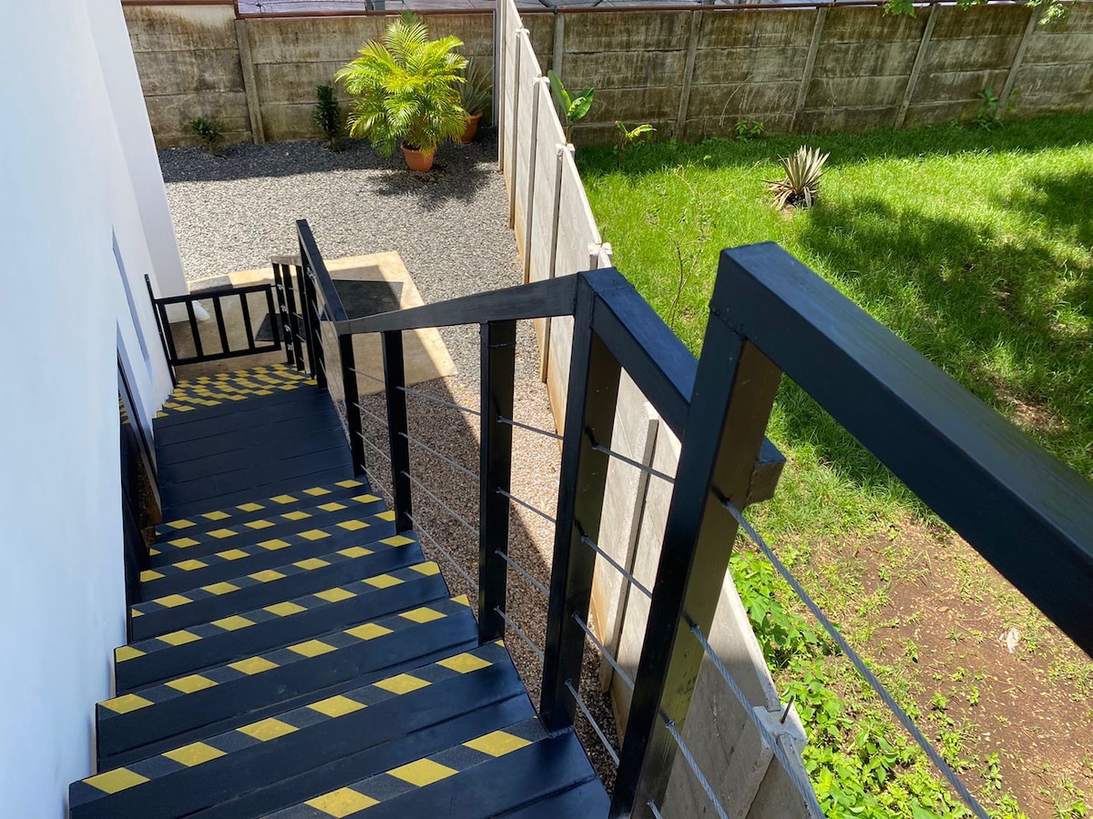 A set of exterior stairs with black steps featuring yellow caution stripes leads down to a gravel path. Lush green grass and potted plants are visible in the garden area, creating a natural backdrop.