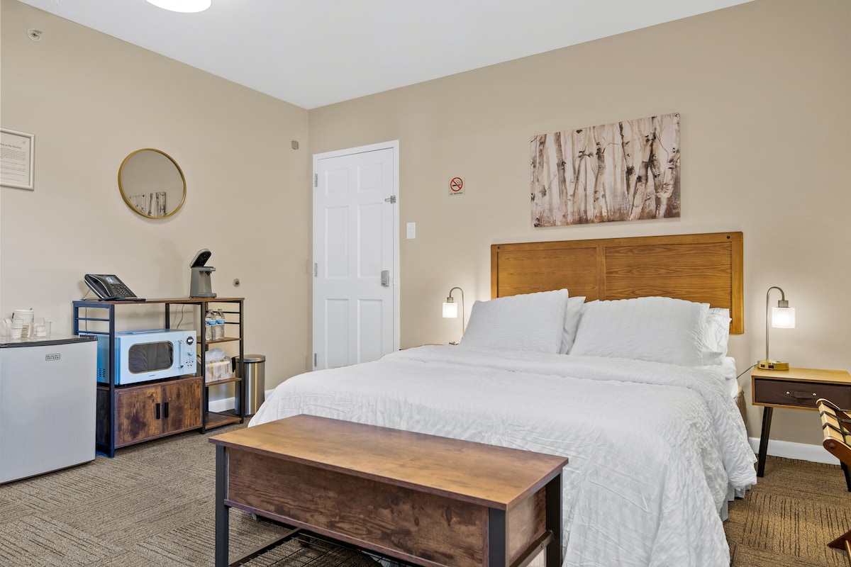 A cozy sleeping area features a Queen bed with light-colored bedding, flanked by bedside lamps. A kitchenette area displays a coffee maker, microwave, and mini refrigerator. A wall art piece adorns the neutral-colored wall, adding a touch of decor to the space.