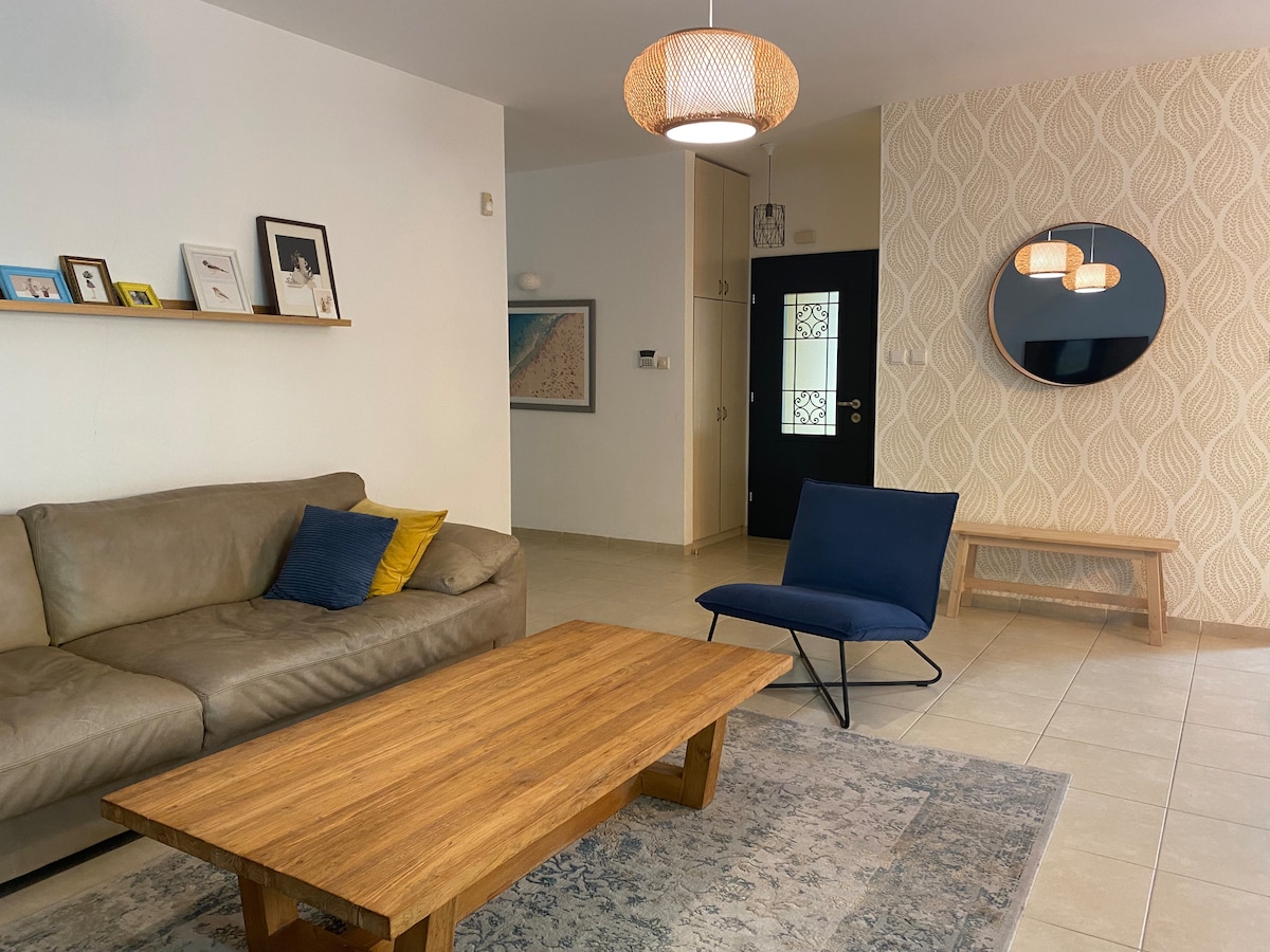 A welcoming living space is presented with a light-toned sofa outfitted with decorative pillows. A wooden coffee table sits in front, paired with a blue chair. Wall decor features framed images, and a round mirror is mounted beside a patterned accent wall.