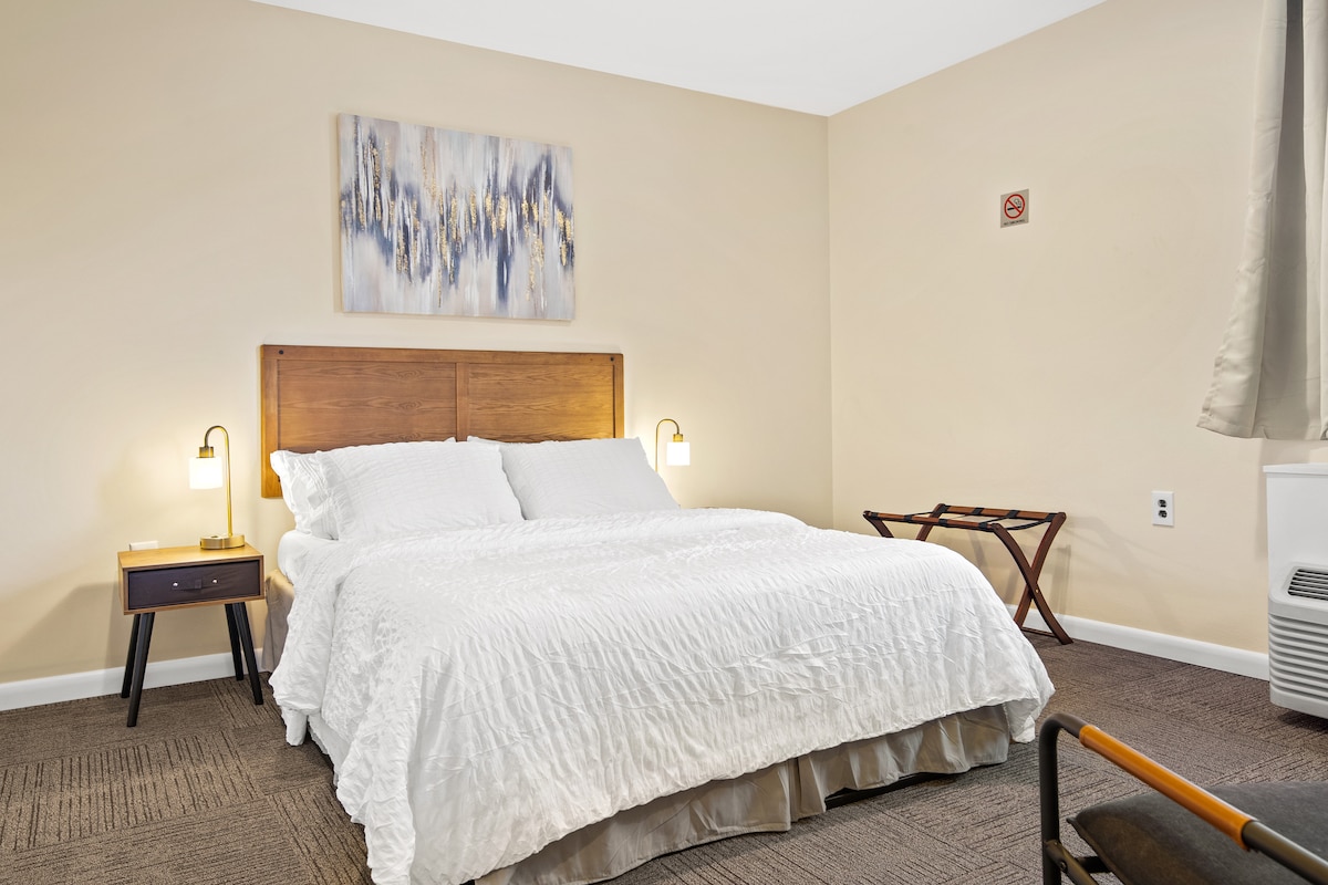 A comfortable bedroom featuring a spacious bed dressed in crisp white linens. Soft lighting is provided by bedside lamps. A small wooden table is positioned beside the bed, while a separate chair is located nearby. Neutral walls create a calming atmosphere.