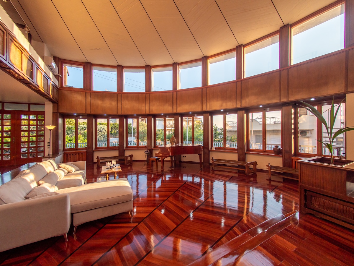 A spacious living room features large wooden frames surrounding multiple windows that fill the area with natural light. Sleek wooden flooring adds warmth, while two comfortable sofas are positioned for relaxation. Various vintage elements, including a piano and dining table, contribute to the room's unique character.