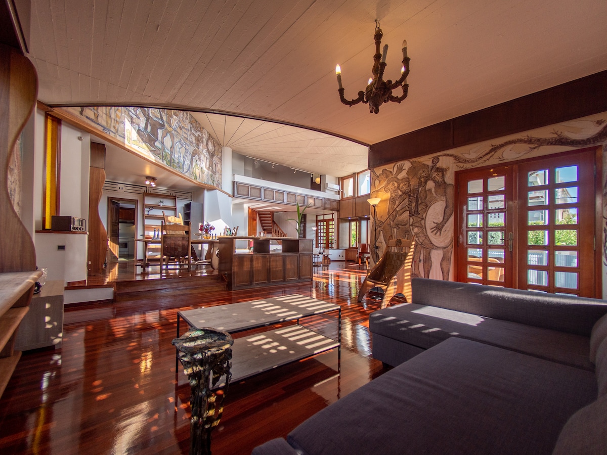 A spacious living area features high ceilings adorned with murals. Large windows allow natural light to accentuate the polished wooden floors. Two comfortable sofas are positioned around a low table, while a dining area and kitchen are visible in the background, blending modern and artistic elements.