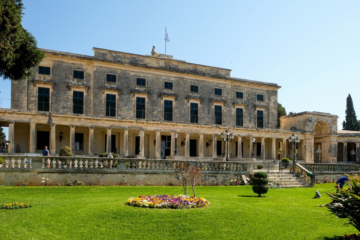 A grand structure showcases classical architecture with multiple columns and large windows. A well-maintained garden features ornamental plants and flower beds in vibrant colors. The building is set against a clear blue sky, providing a picturesque backdrop for the meticulously arranged outdoor space.