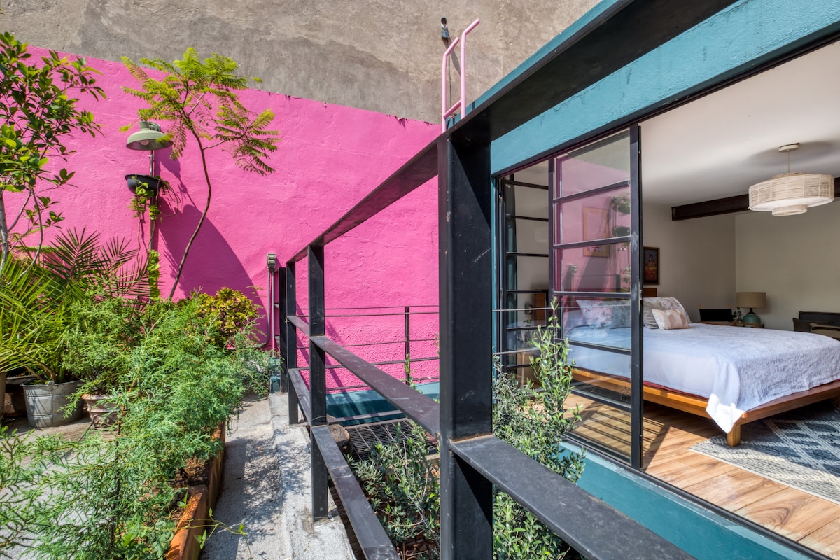 A vibrant outdoor space is framed by a striking pink wall and lush greenery. The view reveals a spacious bedroom through large glass doors, featuring a comfortable bed and modern décor, with natural light illuminating the area.