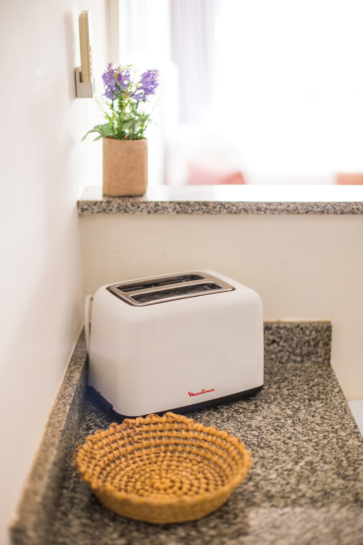 A modern toaster rests on a granite countertop, complemented by a round woven basket. A small vase holding purple flowers adds a touch of color to the kitchen space. Natural light brightens the area from the nearby window.