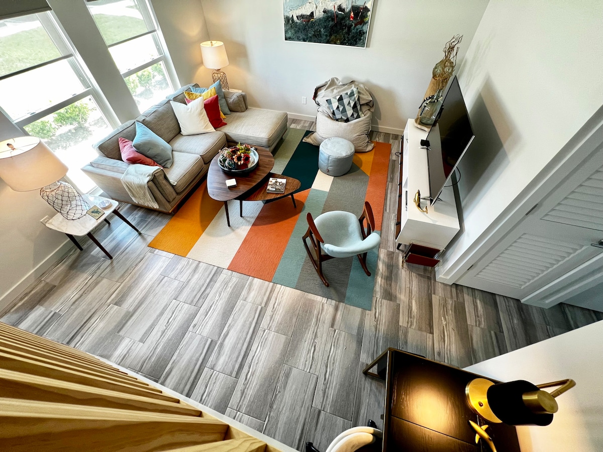 A cozy living area is highlighted by a comfortable sectional sofa adorned with colorful pillows. A round coffee table is surrounded by various seating options, including a light blue chair and a wooden accent chair. Large windows allow natural light to fill the space, enhancing its welcoming design.