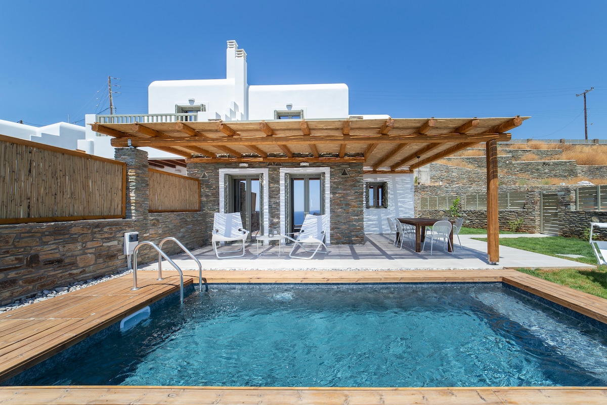 An inviting outdoor space features a rectangular pool surrounded by wooden decking. A shaded pergola offers seating with white lounge chairs and a small table. The backdrop includes stone walls and large glass doors leading into the villa, enhancing the connection between indoor and outdoor areas.