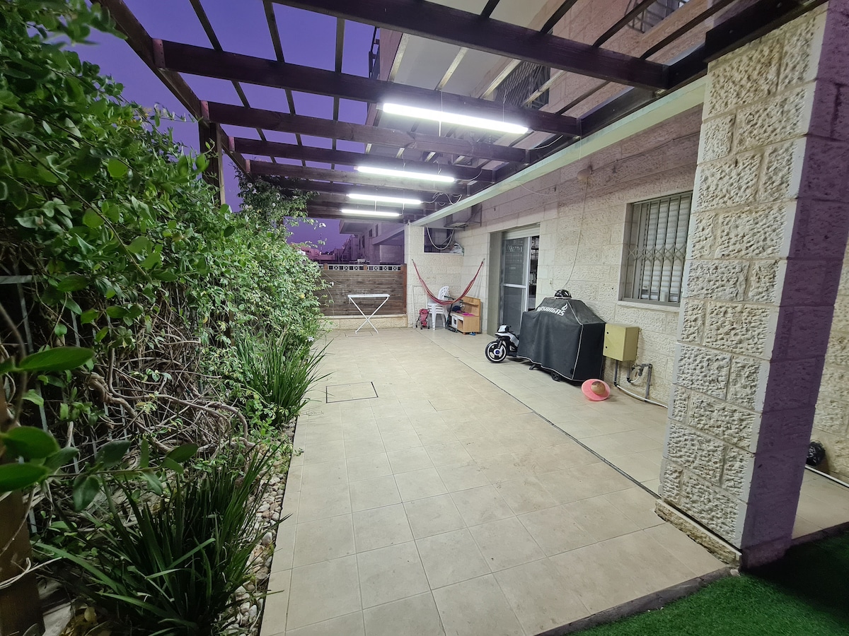 A covered outdoor space features tiled flooring and is bordered by lush greenery. Soft lighting illuminates the area, which includes a grill and a folding table. The layout allows for ample room to relax and enjoy the garden surroundings.