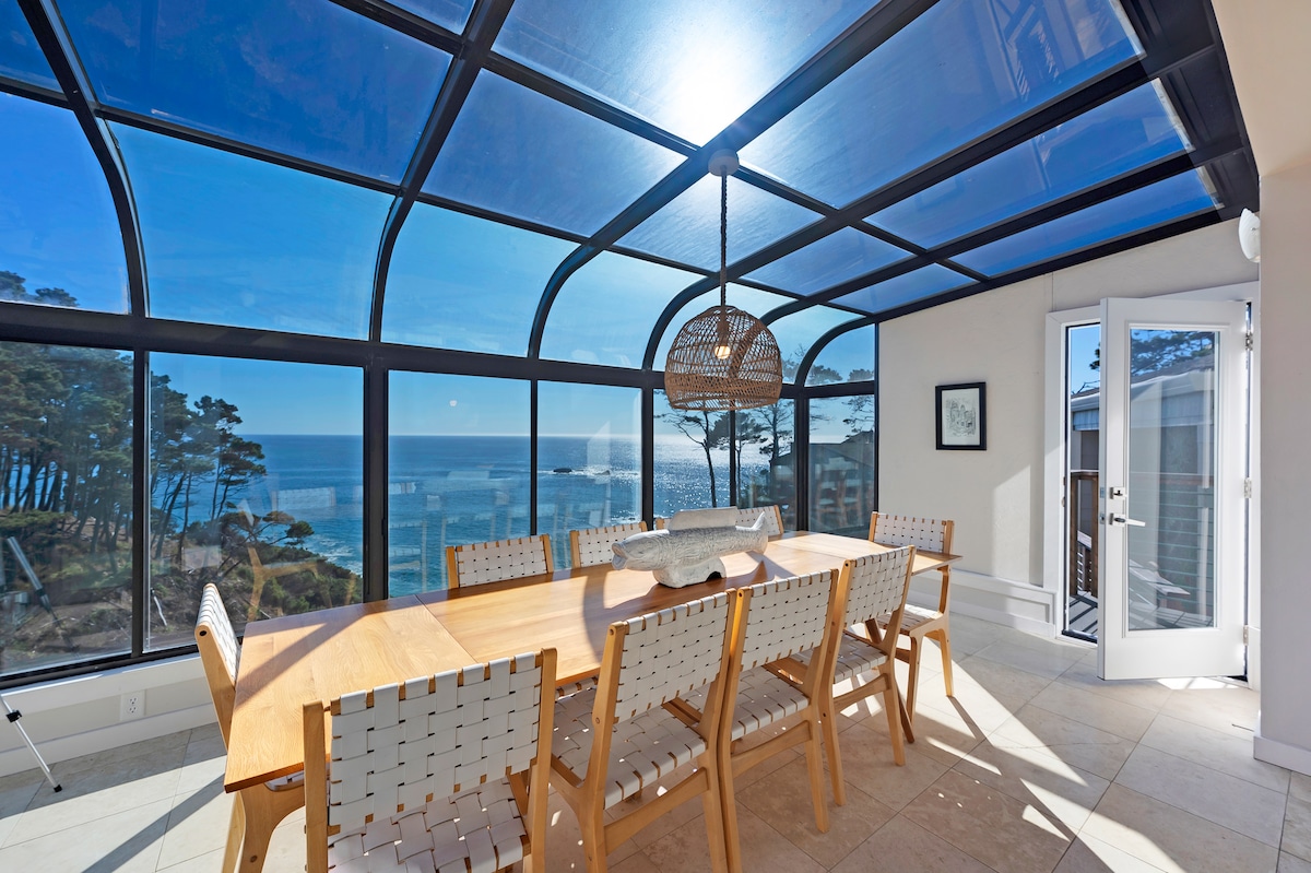 A glass-enclosed dining area is illuminated by natural light, featuring a long wooden table surrounded by ten woven chairs. Expansive ocean views are visible through the transparent walls, and a modern pendant light hangs above the table. An exit door leads to the exterior.