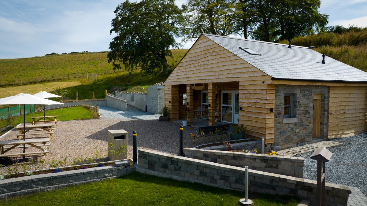 A log-built cabin is showcased amidst a serene landscape, surrounded by grassy hills. A gravel path leads to the entrance, which is framed by a wooden porch. Outdoor seating areas with benches are situated nearby, inviting relaxation in the fresh air.