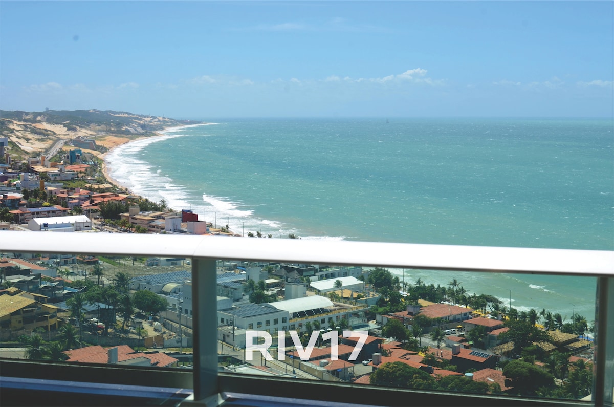 A panoramic view of the sea stretches across the horizon, showcasing the waves gently lapping at the shore. The coastline is lined with residential and commercial buildings, complemented by lush greenery and clear blue skies.