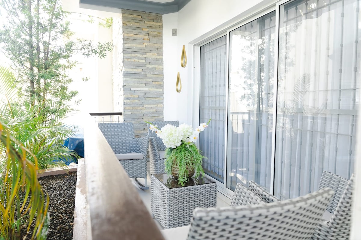 An outdoor balcony space is furnished with woven chairs surrounding a table. A potted plant with white flowers and greenery serves as a focal point, while sunlight filters through sheer window coverings, creating a bright and welcoming atmosphere.