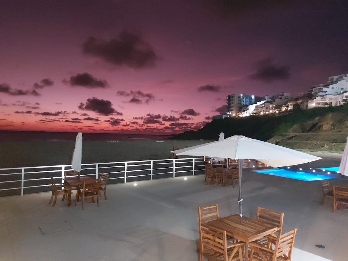 A view of the pool area at sunset features wooden tables surrounded by umbrellas. The horizon showcases a gradient of deep purple and soft pink, reflecting the tranquil seaside atmosphere. The ocean is visible in the background, with buildings illuminated along the coastline.