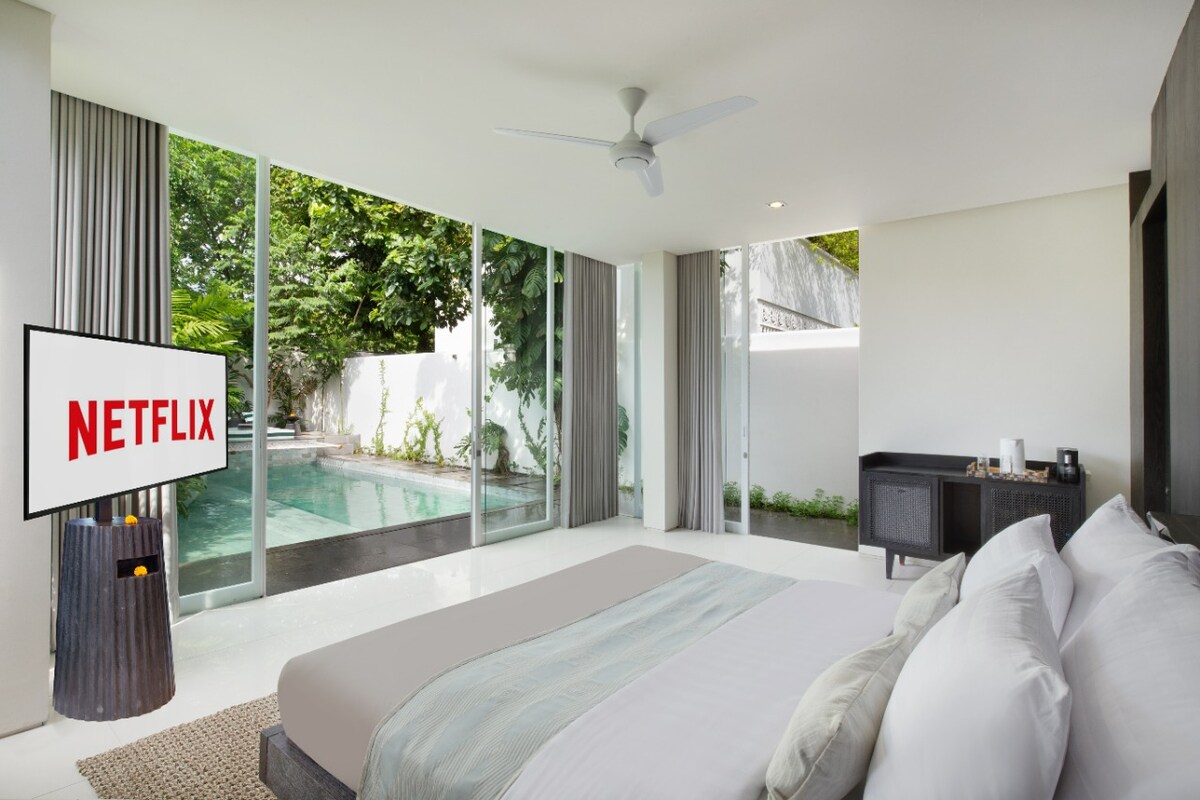 A modern bedroom features a spacious bed with soft linens, complemented by large sliding glass doors that open to a private pool area. A sleek entertainment unit displays a television with streaming capabilities, while minimalist decor enhances the open and airy space.