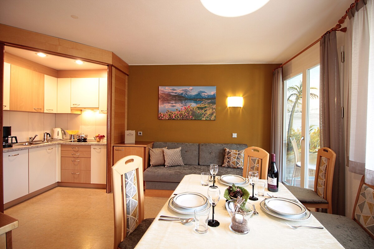 A modern living area features a dining table set for a meal, complemented by stylish chairs. The kitchen, with wood cabinetry and appliances, is in the background. Natural light fills the space, highlighting a decorative piece above the sofa against a warm wall.