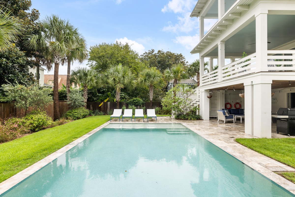 A serene backyard features a refreshing pool surrounded by lush greenery and palm trees. Several loungers are positioned beside the pool, offering a space for relaxation. The spacious patio is visible, with a nearby outdoor grilling area, enhancing outdoor living options.