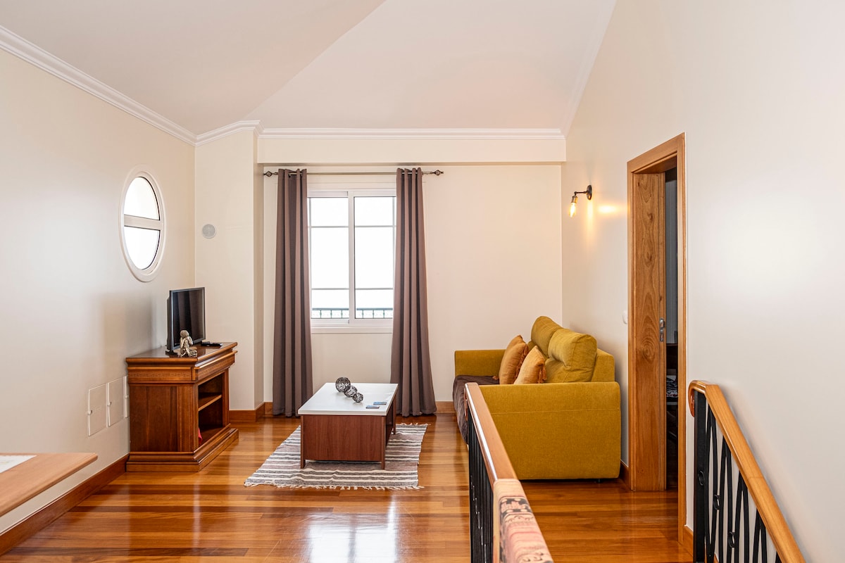 A spacious living area features large windows allowing natural light to fill the room. A comfortable sofa and a wooden coffee table are positioned on a hardwood floor, complemented by a soft rug. A television stands on a wooden cabinet, enhancing the cozy atmosphere.