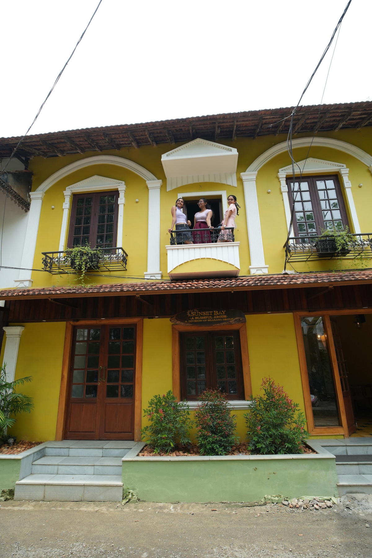 The charming exterior of the building showcases bright yellow walls, elegant white arches, and intricate wooden doors. Balconies adorned with potted plants add a touch of greenery. A well-maintained pathway leads to the entrance, enhancing the inviting appearance of this vibrant structure.