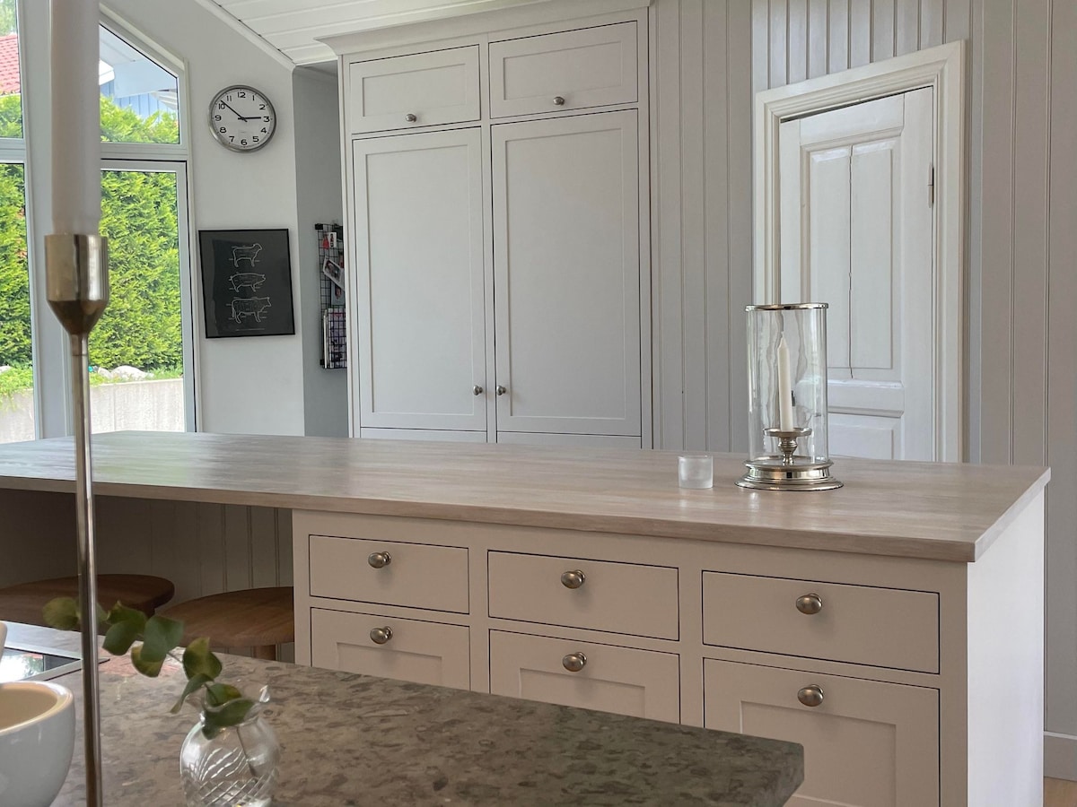 A modern kitchen showcases an island with a light wooden countertop and multiple storage drawers. The cabinetry is designed with a neutral tone, complemented by bar stools for seating. A clock is mounted on the wall, and natural light filters through large windows.