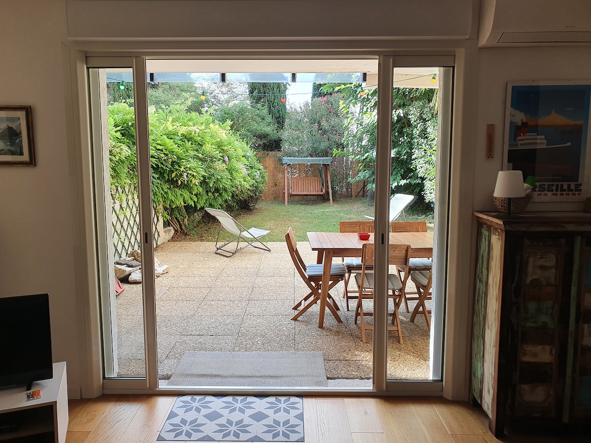 A clear view is presented through sliding glass doors, revealing a private outdoor space. A wooden dining table with chairs is positioned on a stone patio, surrounded by lush greenery and a relaxation area with loungers. The garden features a wooden swing set in the background.