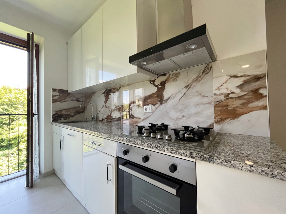 The kitchen features modern white cabinetry and a stylish marble backsplash. A four-burner gas stove and an oven are present on the granite countertop. Natural light enters through the window, offering a view of greenery outside.