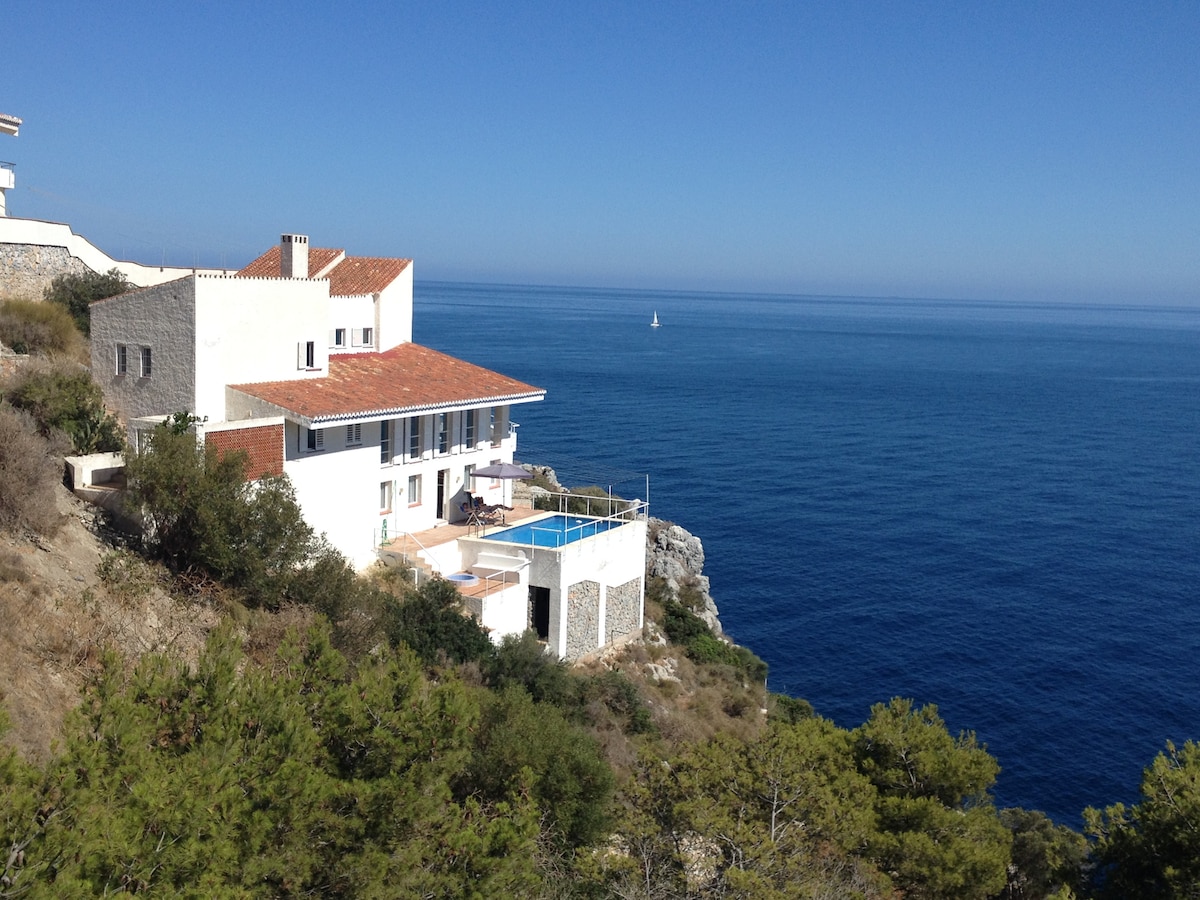 A view of the modern house perched on a rocky cliff overlooking the sea. The building features a spacious terrace with a private pool, surrounded by natural vegetation. The expansive ocean stretches into the horizon, where a sailboat can be seen in the distance.
