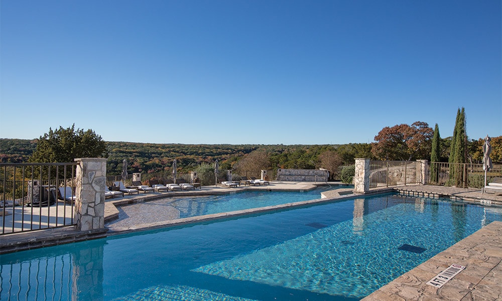 An outdoor swimming pool is set against a scenic hillside backdrop. The pool features a stone border and spacious deck area with lounge chairs. Lush greenery and trees can be seen in the distance, under a clear blue sky.