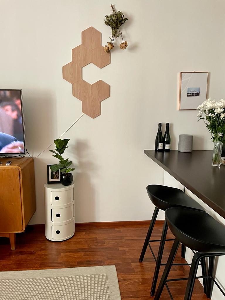 A modern living area features a wooden wall decoration with florals above a sleek bar table. Two black chairs are positioned at the bar. A television stands beside a vintage cabinet, and a small plant is placed on a cylindrical storage unit.