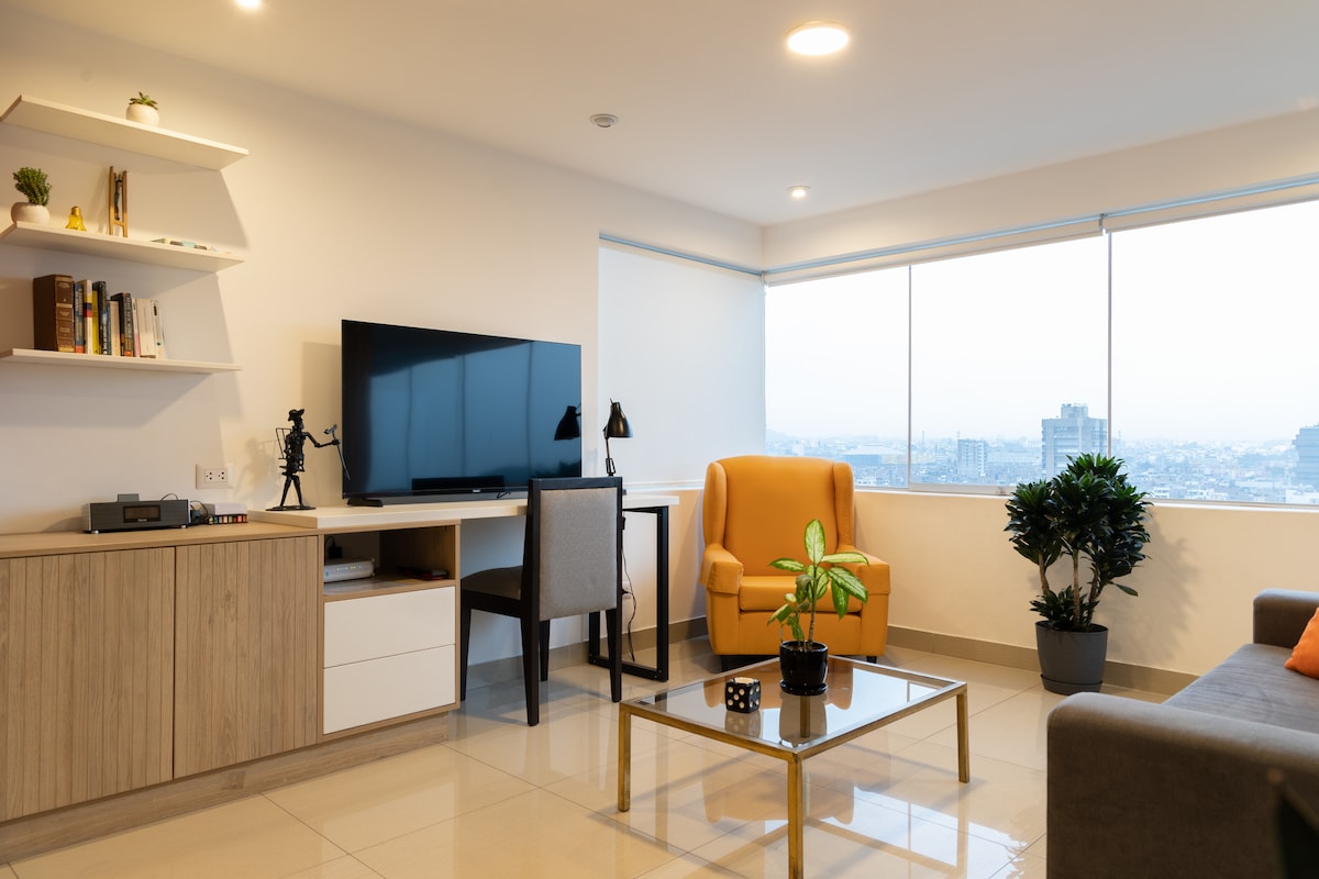 A modern living area is displayed, featuring a spacious sofa and a bright yellow armchair. A glass coffee table sits centered, complemented by a small potted plant. A large television is mounted on the wall, with a workspace including a desk and chair located nearby. Expansive windows reveal panoramic city views.