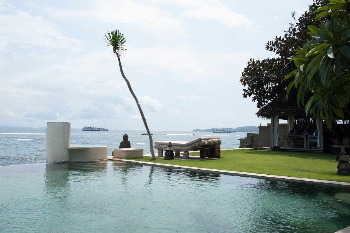 An infinity pool reflects the sky and ocean, seamlessly extending toward the horizon. A lone palm tree rises over the water's edge, offering subtle shade. Nearby, traditional seating is visible along the manicured lawn, while distant boats gently navigate the ocean.