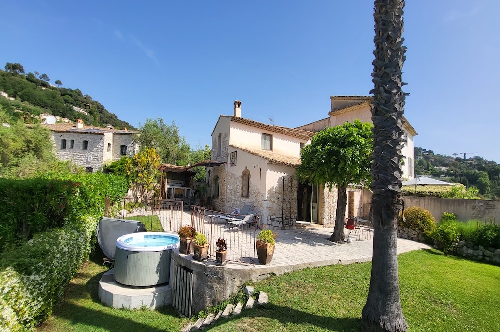 A private garden is showcased with well-maintained grass and greenery. A jacuzzi is positioned against a stone wall, surrounded by sun loungers. The villa, featuring a stone façade and a terracotta roof, is visible in the background under a clear blue sky.