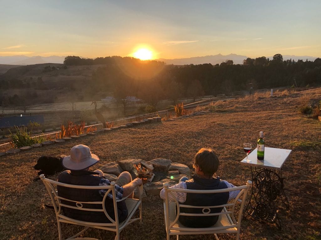 Two individuals are seated on metal chairs facing a picturesque sunset over rolling hills. A small stone table holds drinks, while an outdoor fire pit is nearby, enhancing the serene atmosphere of the rural landscape. The warm hues of sunset illuminate the scene.